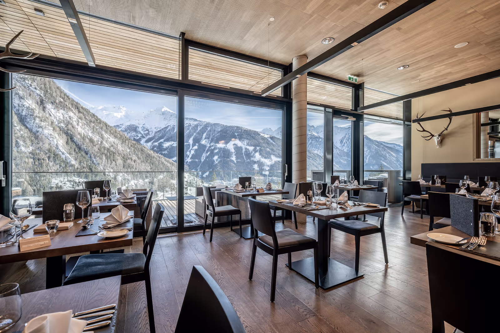 Tables are elegantly set with glasses and napkins in a stylish restaurant overlooking snow-capped mountains through large glass windows, creating a serene and upscale dining atmosphere.