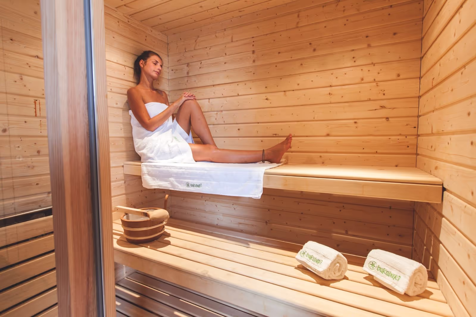 A person wrapped in a towel relaxes on a wooden sauna bench. Nearby, a bucket is placed along with towels labeled "Isokinetik," creating a calming spa environment.