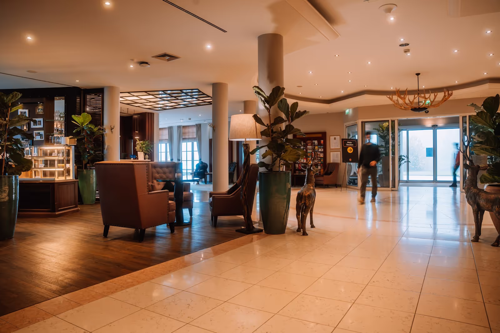 Lobby with armchairs and a coffee table under warm lighting, surrounded by potted plants and deer sculptures; two people walk across the polished floor toward glass exit doors.