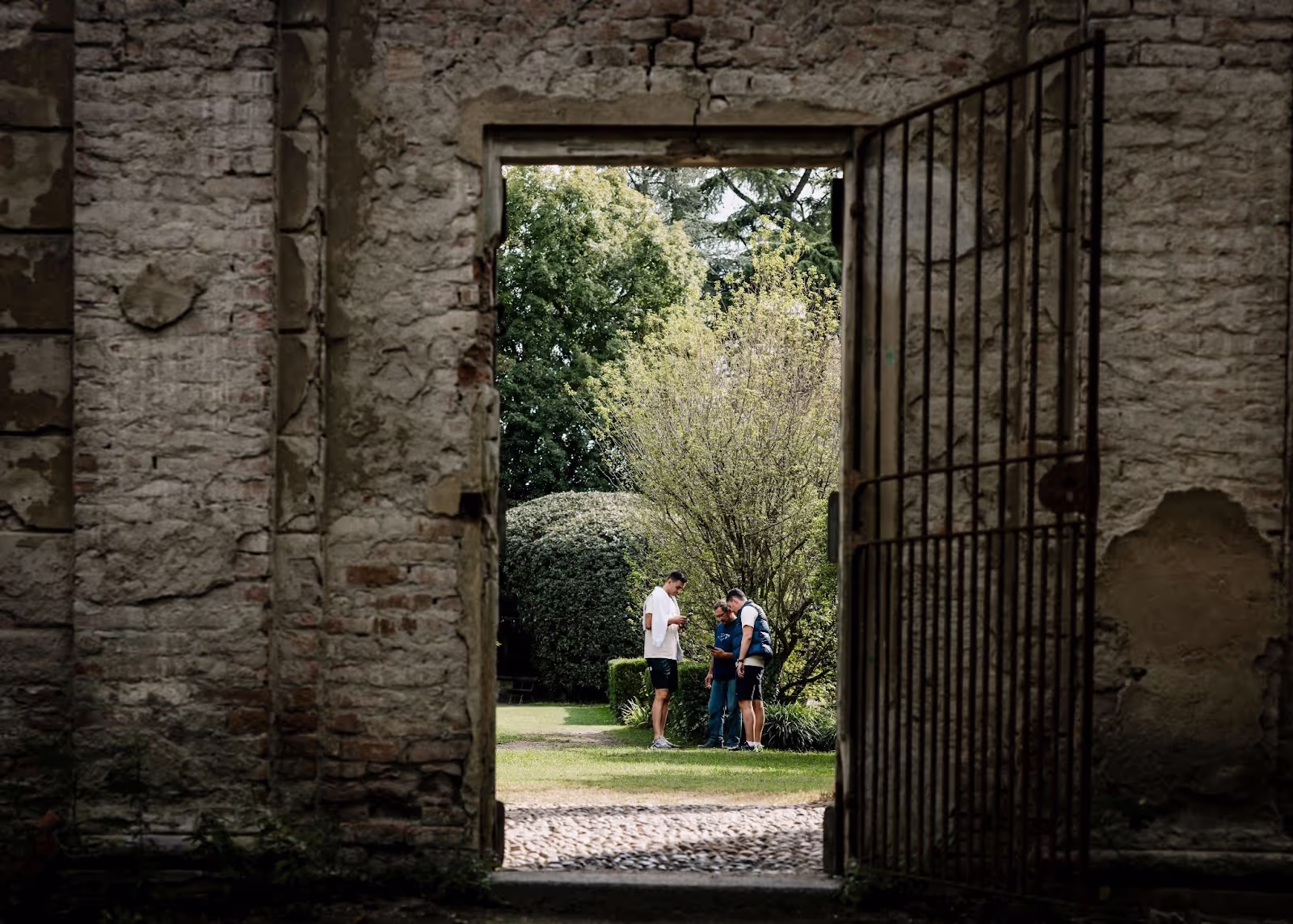 People conversing stand in a lush garden, visible through a weathered stone doorway with an open metal gate. Surrounding greenery includes trees and shrubs, evoking a serene, rustic ambiance.