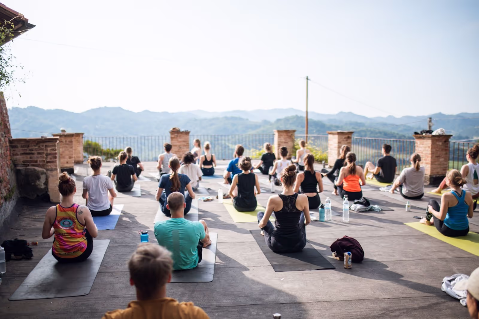 People are seated on yoga mats, meditating outdoors on a terrace. Surrounding them are scenic, distant mountains and a clear sky, creating a peaceful environment.