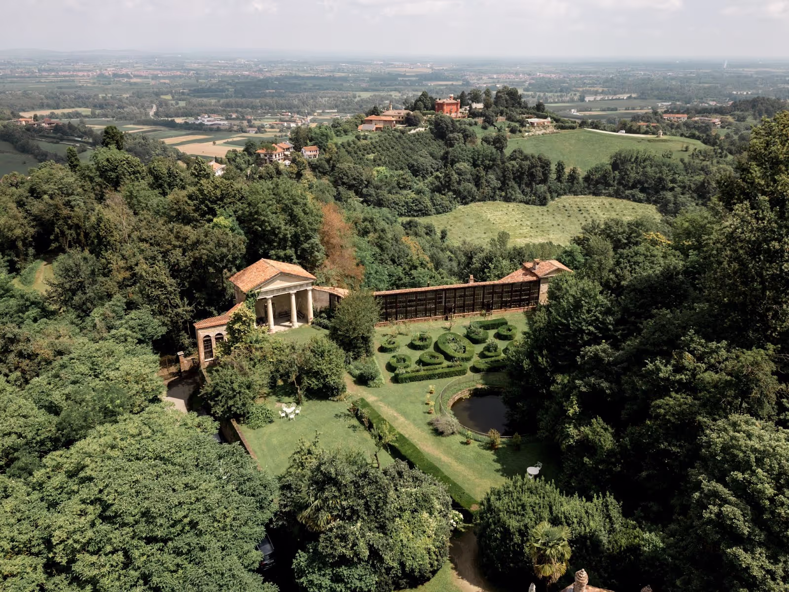 A neoclassical pavilion with columns overlooks manicured gardens featuring topiary and a pond, set amidst lush greenery and rolling hills extending into the distant landscape.