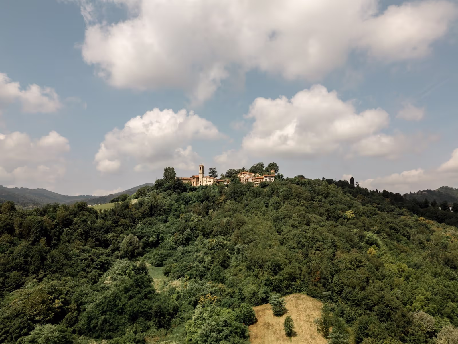 A hilltop villa with a tower stands amidst dense green forest, under a cloudy blue sky. Hills surround the area, enhancing the picturesque, serene landscape.