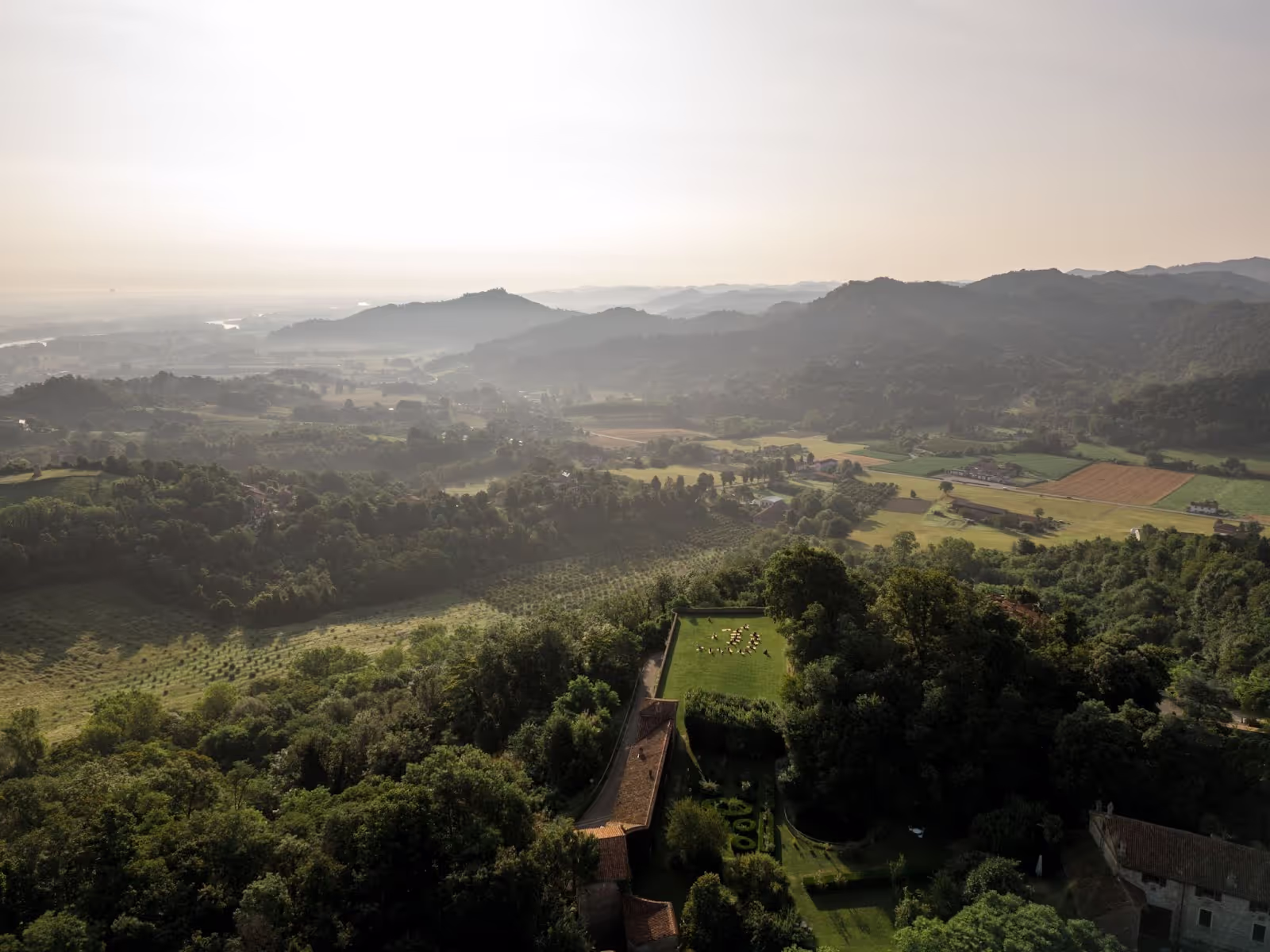 Aerial view of a lush hillside, with a large grassy area containing arranged yellow chairs, bordered by dense trees; distant rolling hills and fields under a hazy sky create an expansive landscape.
