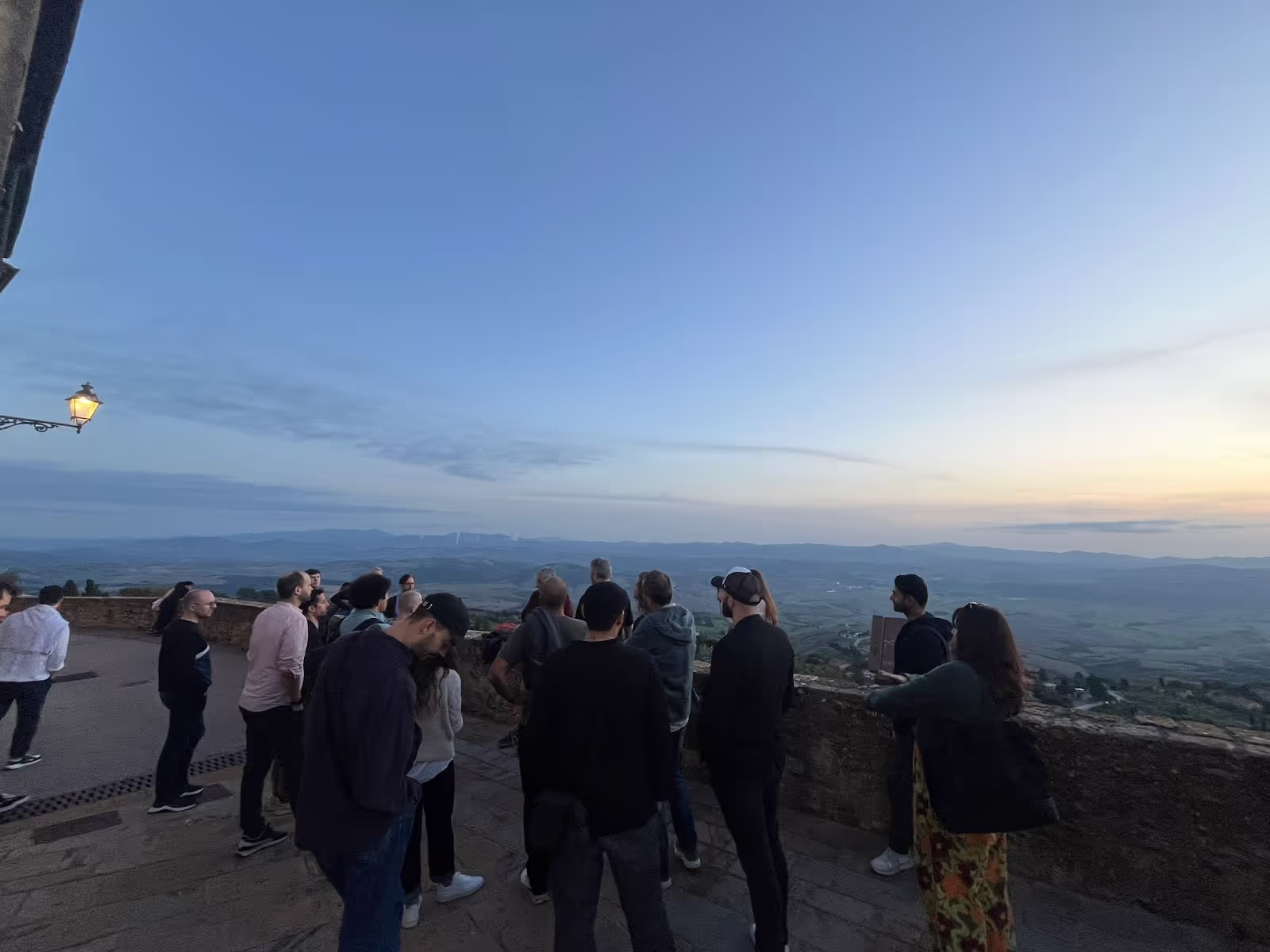 People standing in a group, gazing at a distant scenic landscape during sunrise or sunset, with rolling hills and soft light illuminating the sky, near an outdoor stone wall.