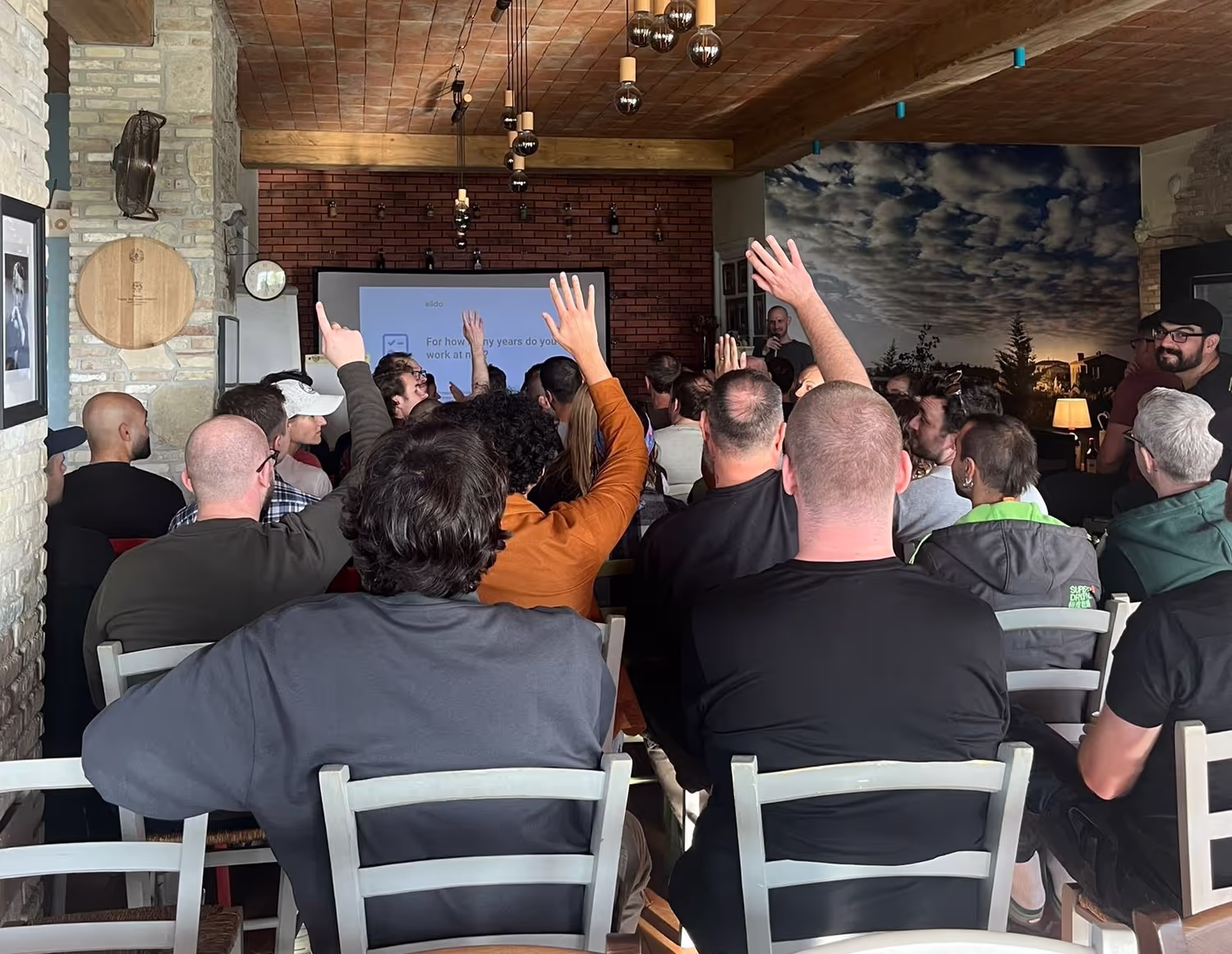 Audience members raising hands during a presentation in a cozy room with a brick wall and scenic wallpaper. A slide on the screen reads, "For how many years do you work at..."