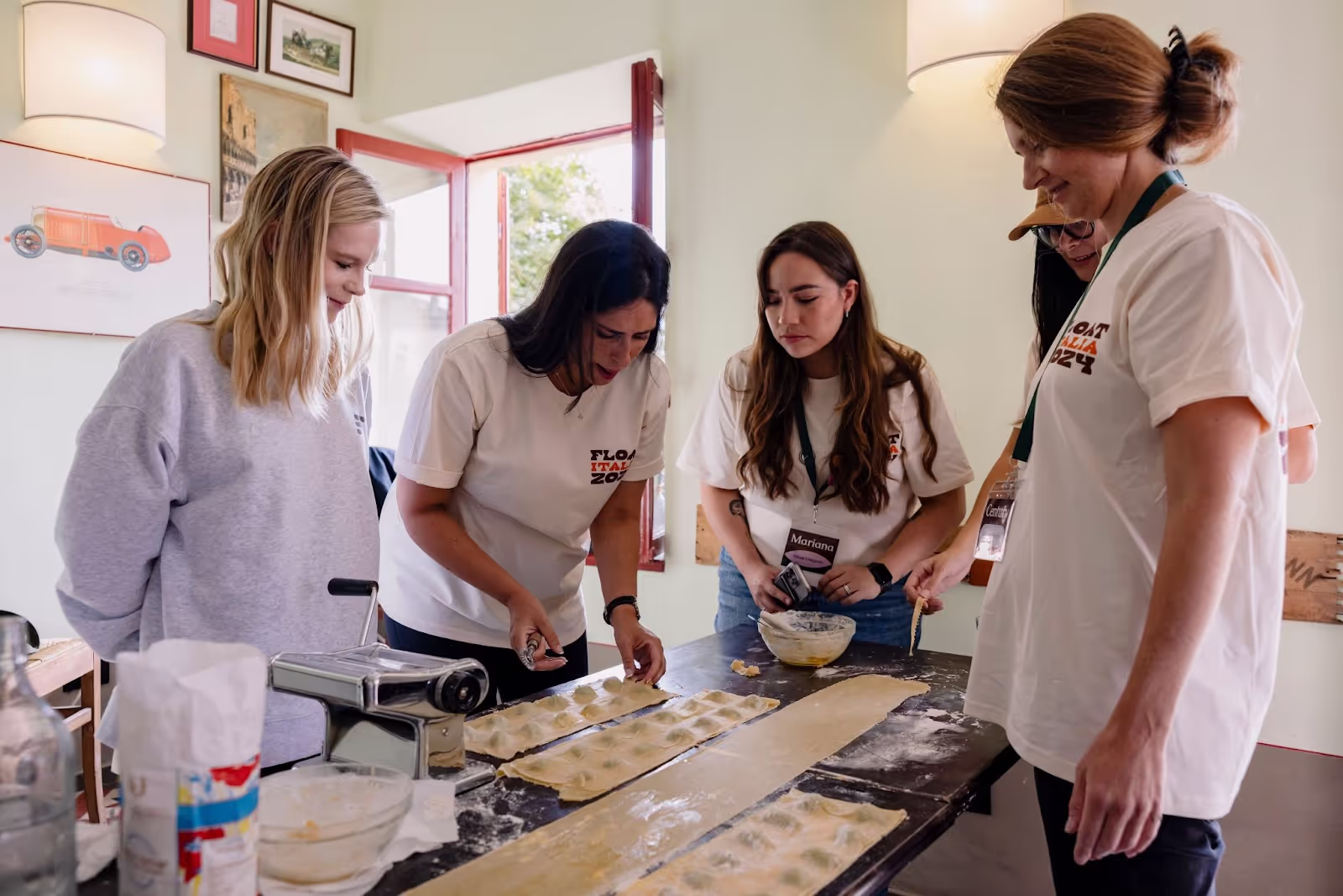 People gather around a table making pasta using a machine and flour, in a well-lit room with framed art and an open window. Shirts read "FLOAT ITALY 2024".