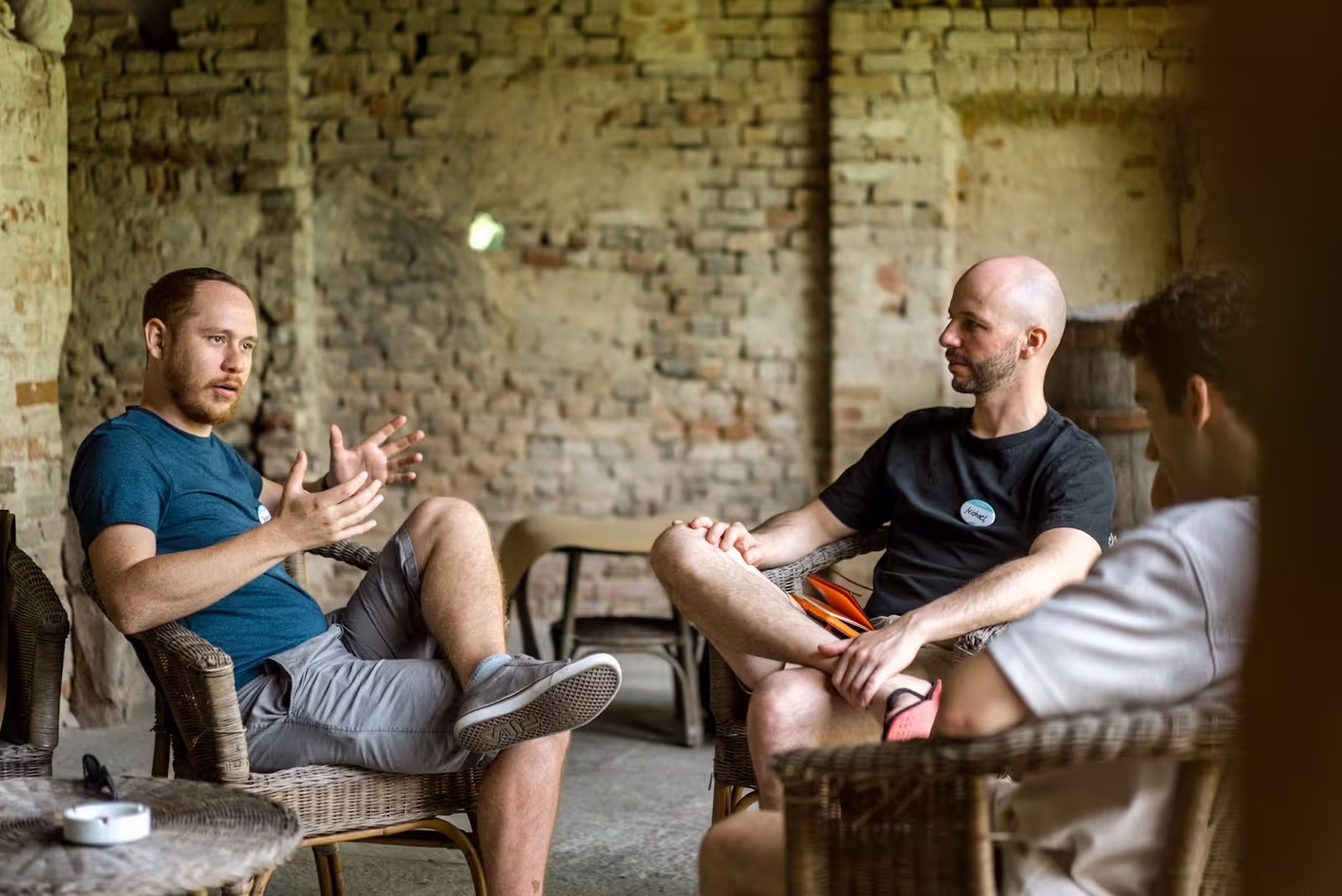 Three people sit and converse in wicker chairs against a rustic brick wall. One gestures expressively, engaging the others, creating an informal, relaxed atmosphere.
