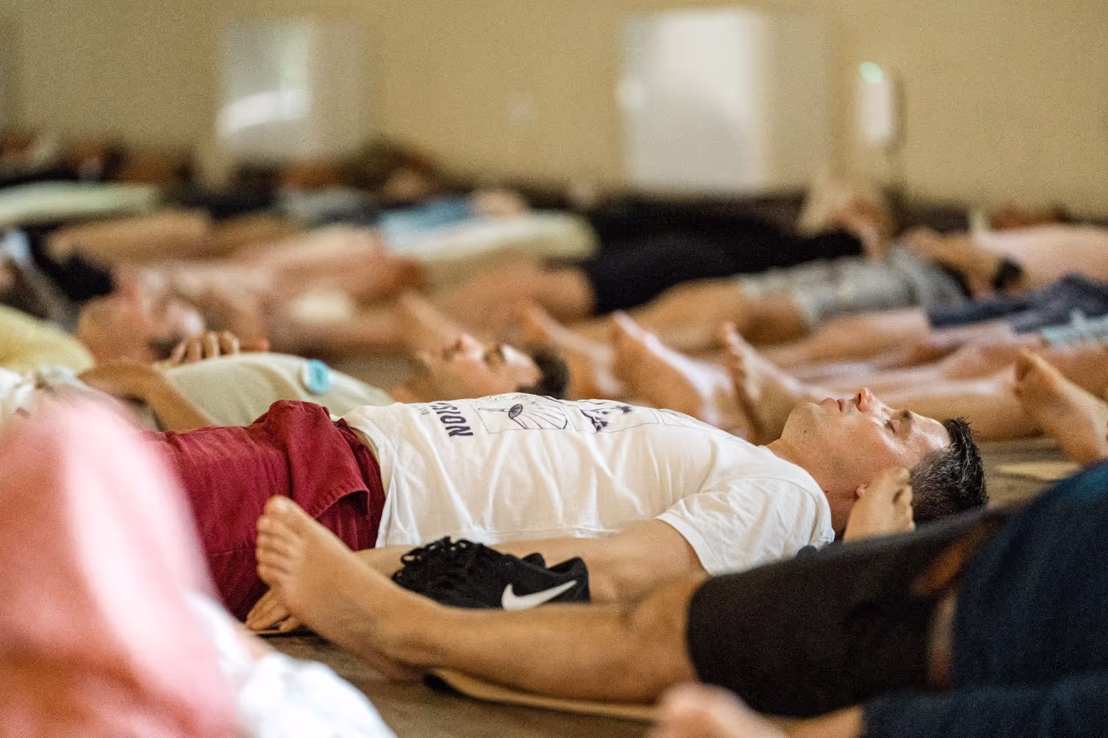People are lying on yoga mats, resting in a relaxation pose in a calm indoor setting. A man in the foreground wears a white shirt with an indistinct print and red shorts.