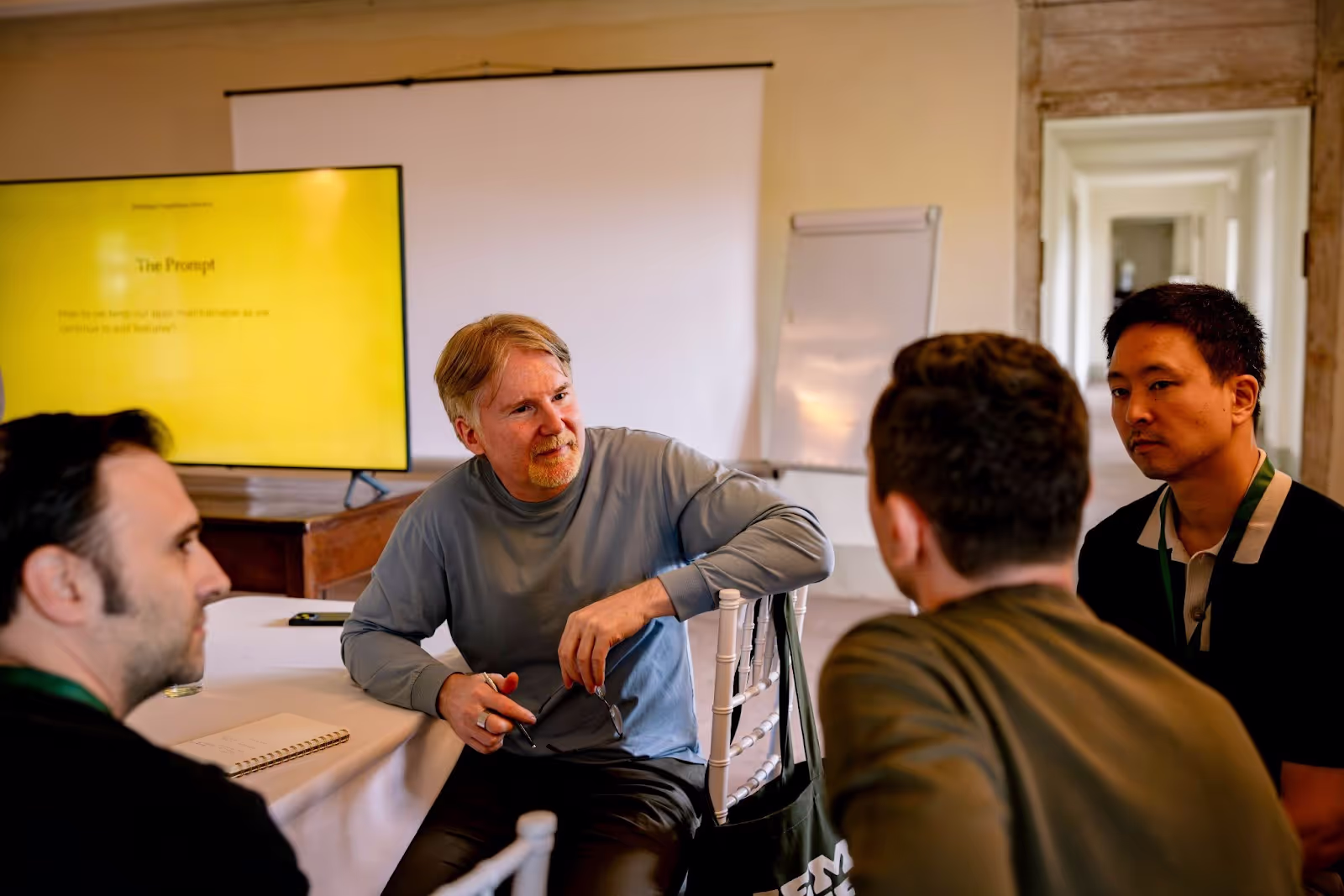 Four people are engaged in a discussion around a table in a room with a monitor displaying text: “The Prompt: How do we keep our apps maintainable as we continue to add features?”