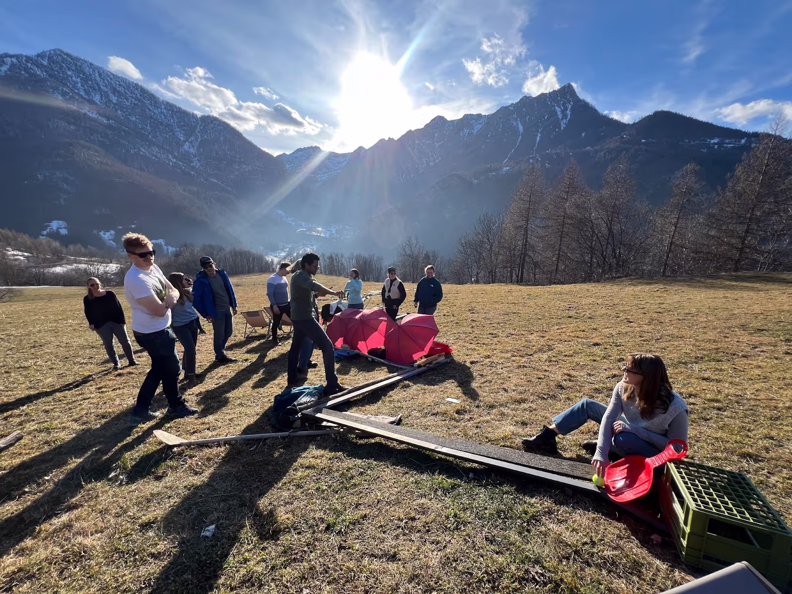 People are gathered outdoors on a grassy field, enjoying sunlight and mountainous scenery in the background. A person sits on the ground near makeshift sledding equipment, while others stand and chat.