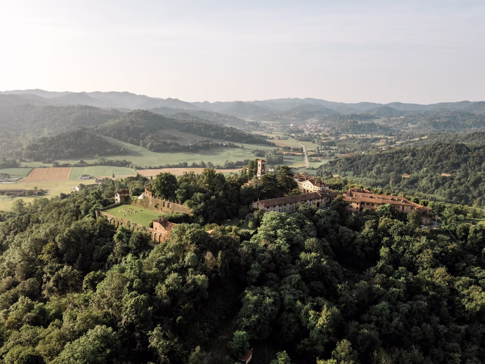 A large, historic complex of buildings with red-tiled roofs sits atop a forested hill, surrounded by rolling green hills and fields under a clear sky.