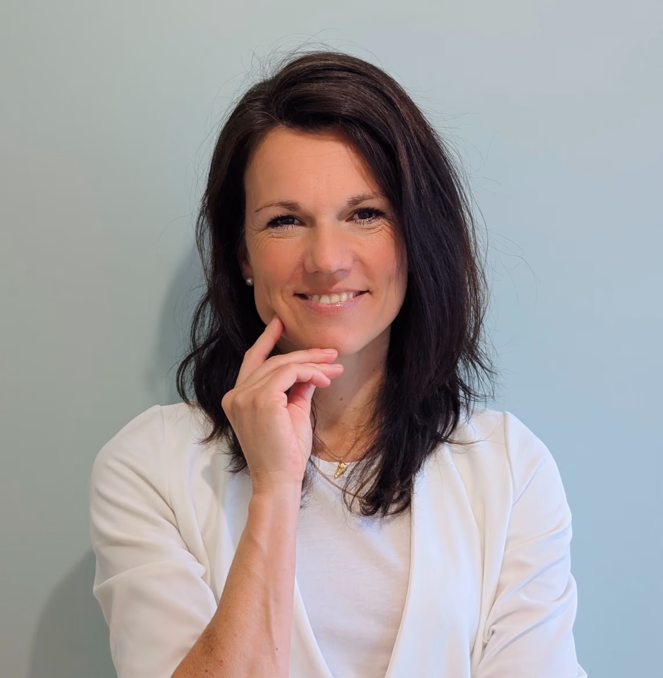 Smiling woman with dark shoulder-length hair wearing a white top posed against a light blue background.