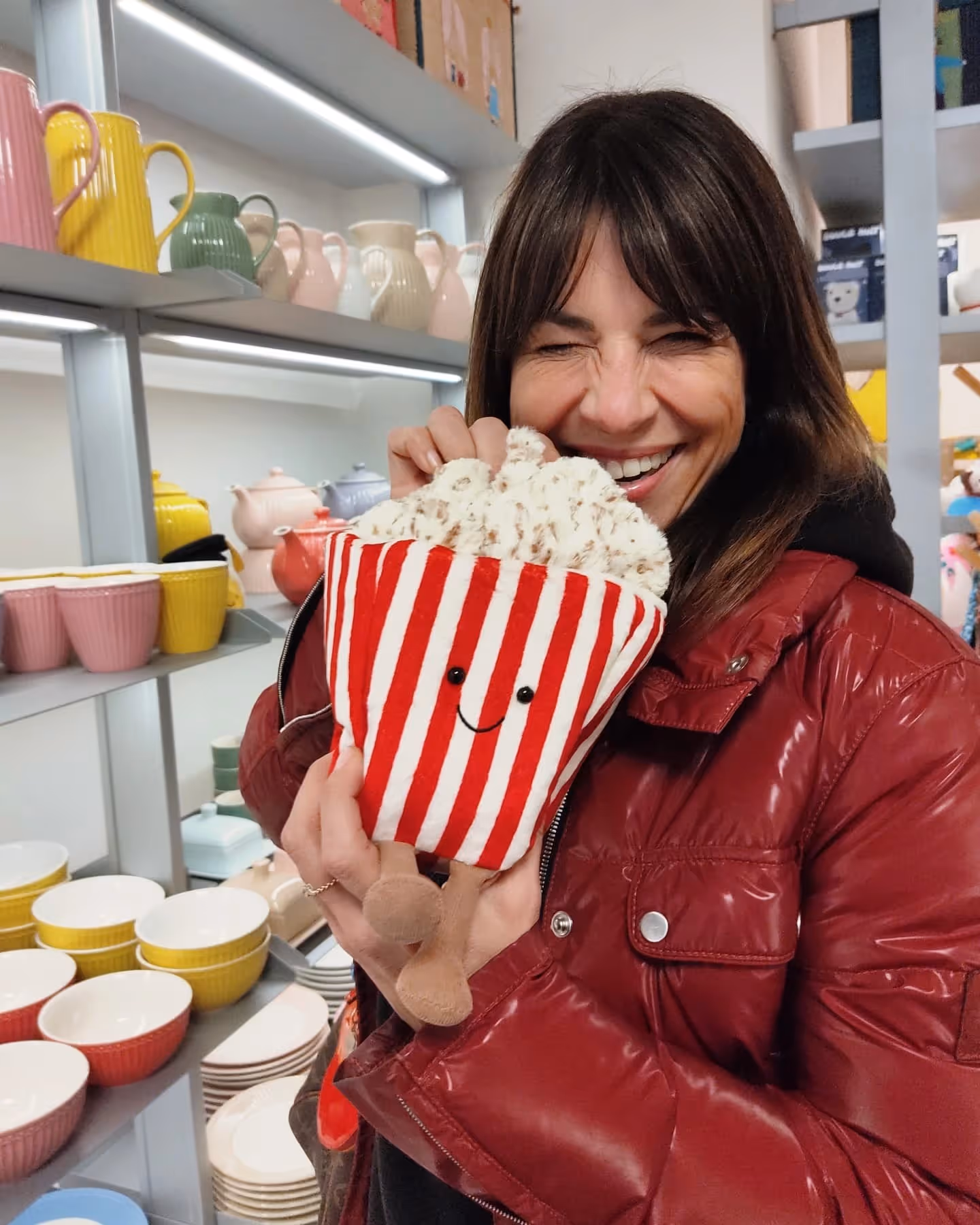Frau in rotem Mantel lächelt und hält ein Plüschtier in Form von Popcorn mit Gesicht.