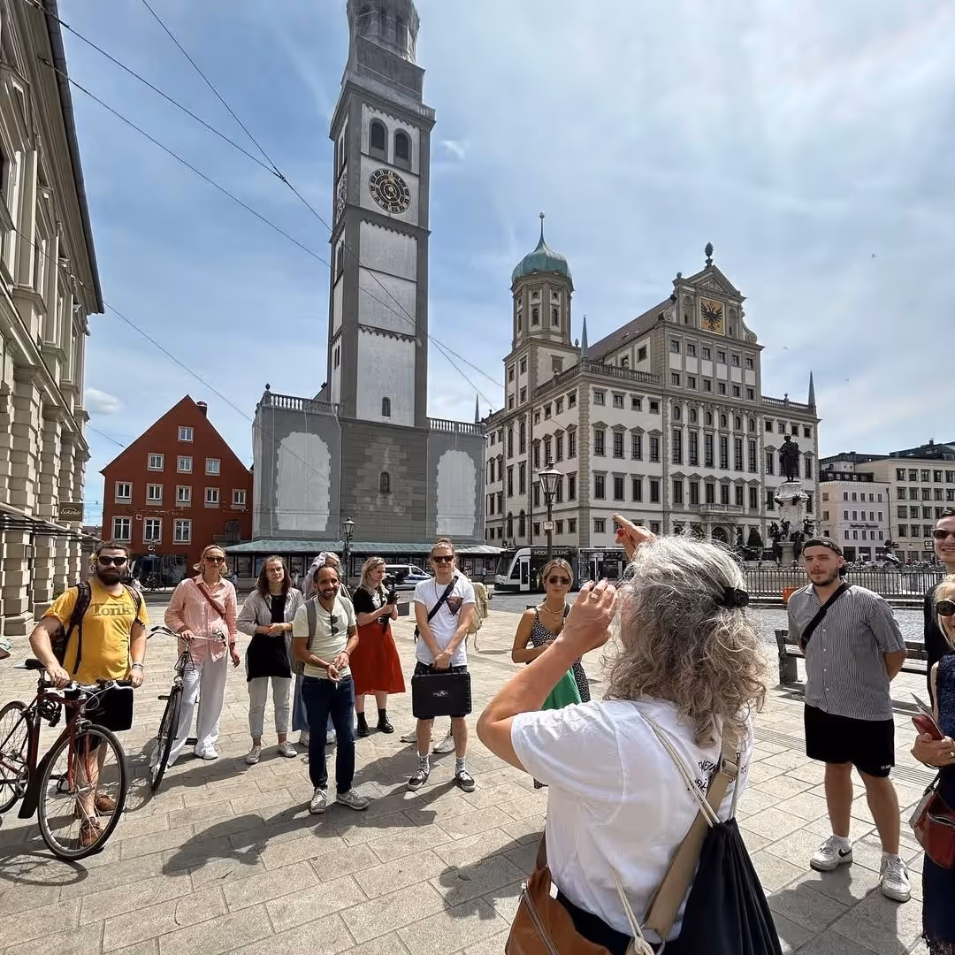 Führerin zeigt Gruppe von Touristen auf historische Gebäude in einer sonnigen Stadt.
