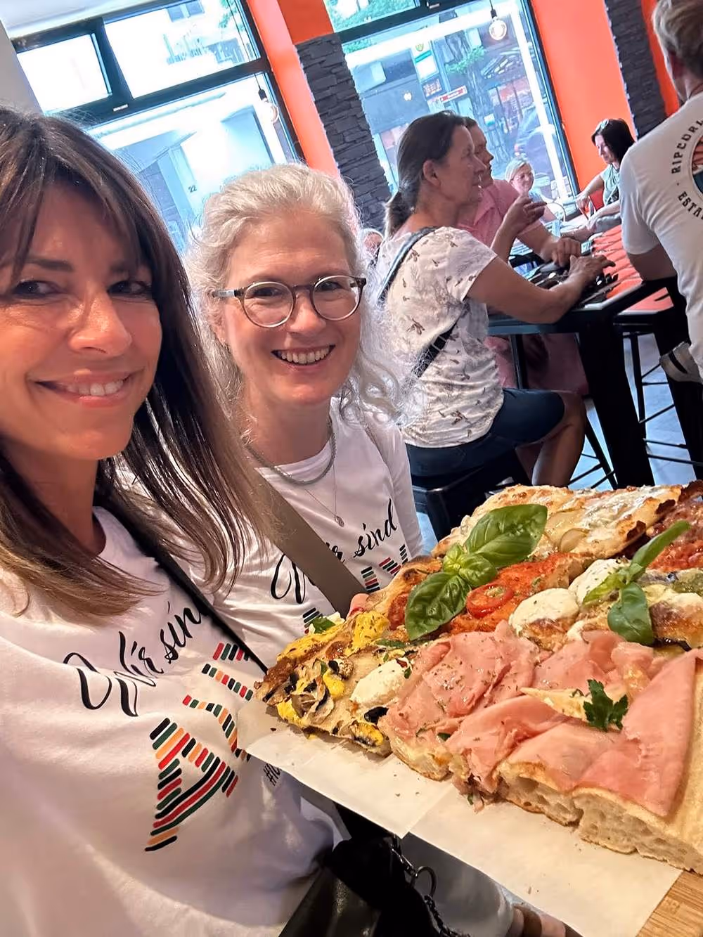 Zwei Frauen halten ein großes Blech mit verschiedenen Pizzastücken und lächeln in die Kamera.