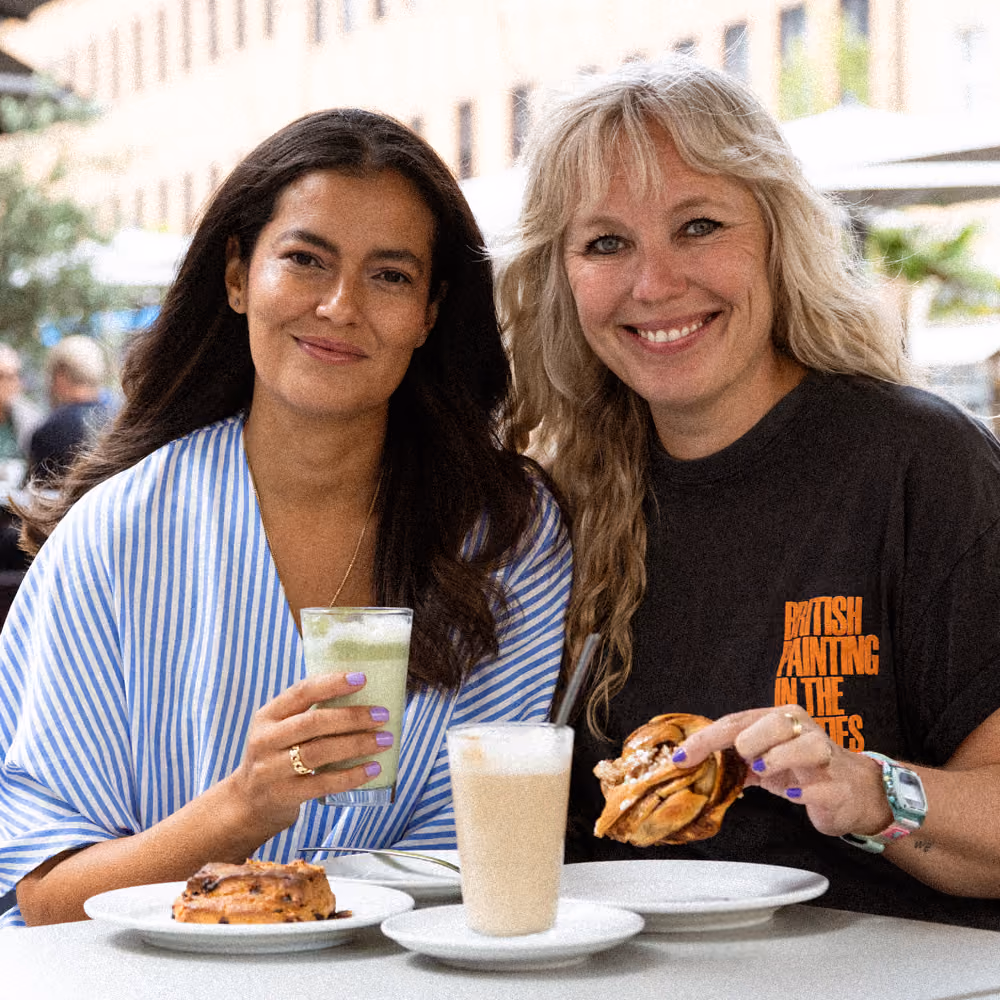 Zwei Frauen sitzen draußen, eine hält einen Matcha Latte, die andere isst Gebäck und hat Kaffee.