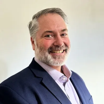 A man wearing a blue jacket and a beard smiles for the camera.