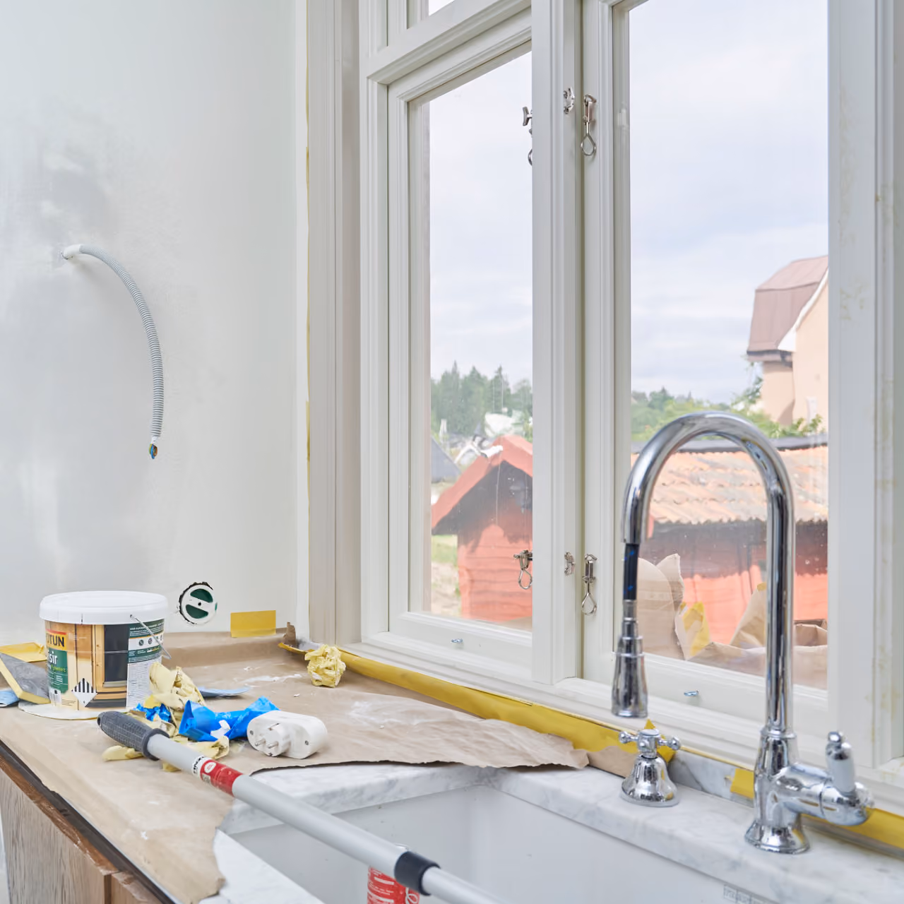 Nyt kök under renovering med arbetsbänk, kran, målarsaker och utsikt mot röda stugor genom fönster.