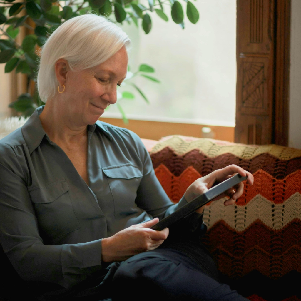 An elderly white woman with short white hair wearing a blue shirt cozies up on the couch holding a picture frame and smiling.