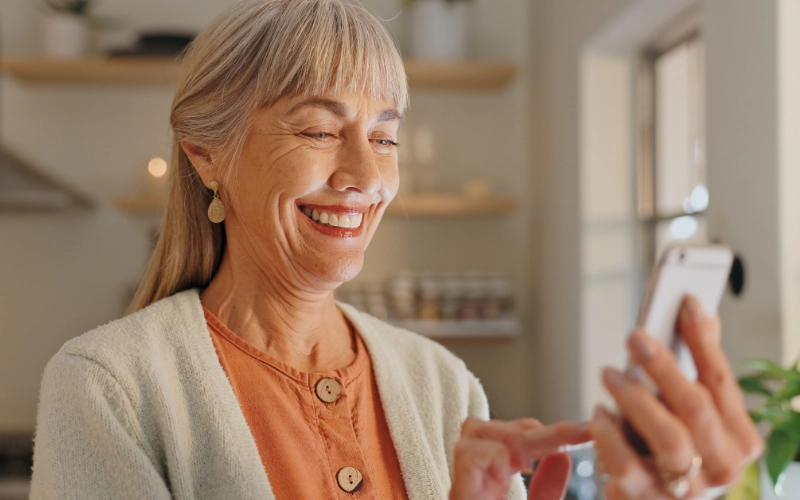 An elderly white woman with long silver hair pulled back wearing an orange shirt and holding her phone as she touches the screen and smiles.