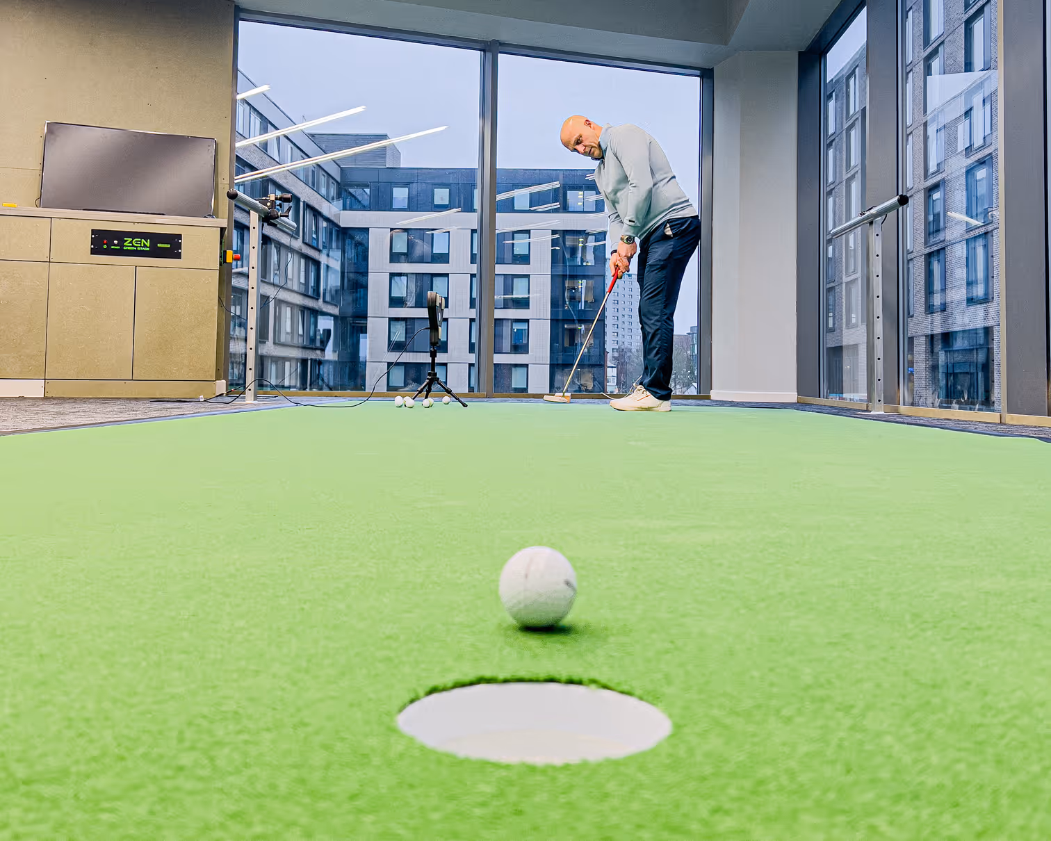 Person preparing to hit a golf ball indoors with a golf bag labeled 'FRINGEGOLF' on artificial turf and a golf simulation screen in the background.
