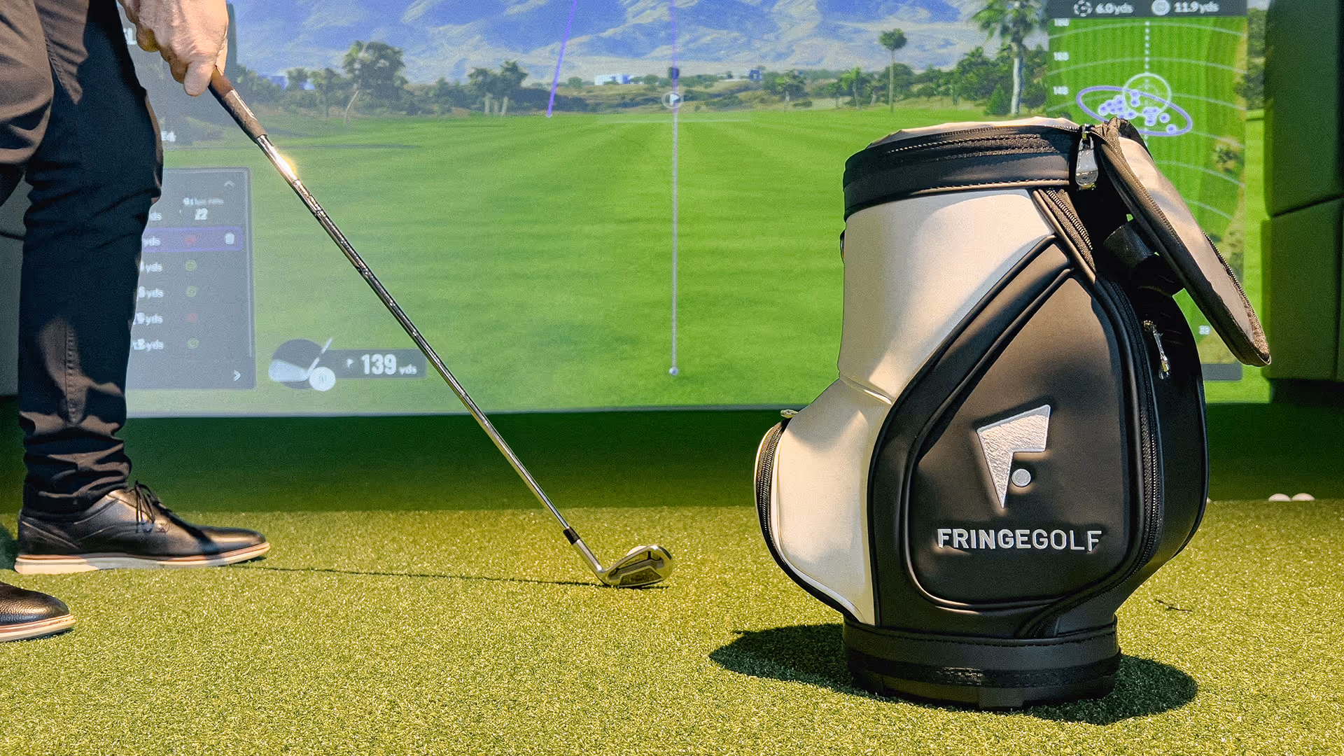 Person preparing to hit a golf ball indoors with a golf bag labeled 'FRINGEGOLF' on artificial turf and a golf simulation screen in the background.