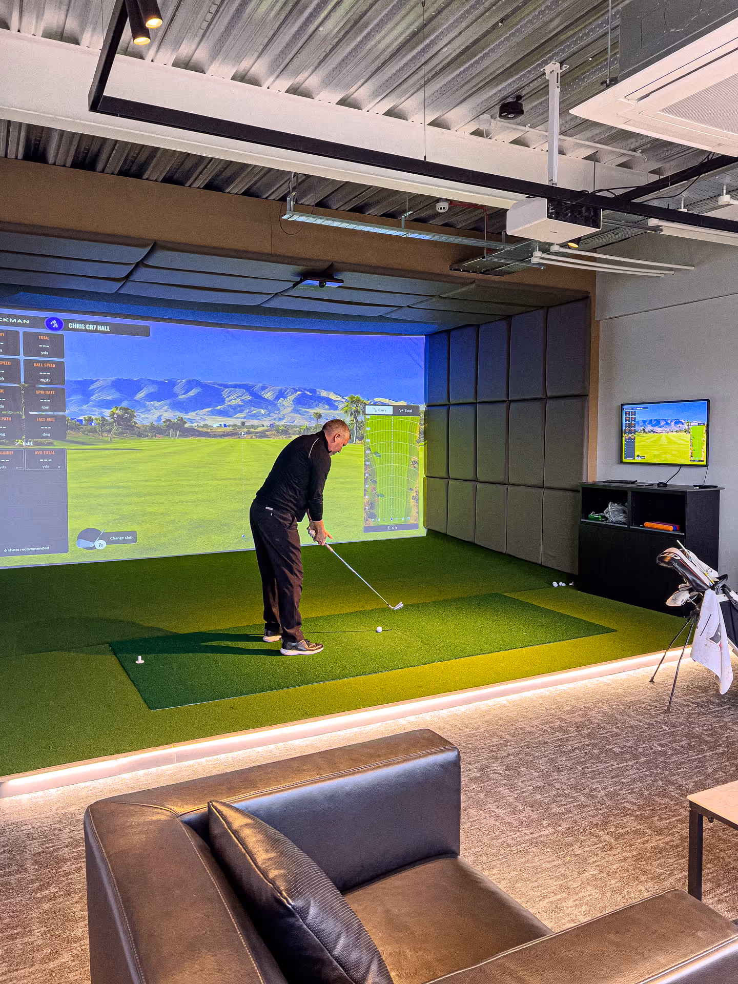 Person preparing to hit a golf ball indoors with a golf bag labeled 'FRINGEGOLF' on artificial turf and a golf simulation screen in the background.