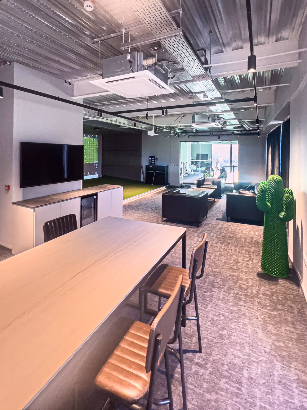 Person preparing to hit a golf ball indoors with a golf bag labeled 'FRINGEGOLF' on artificial turf and a golf simulation screen in the background.