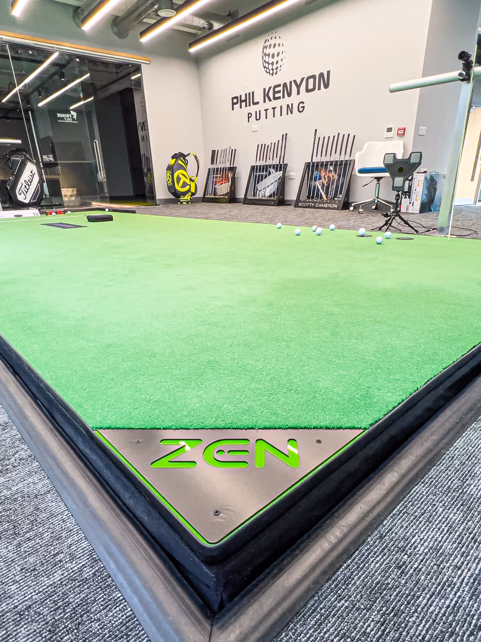 Person preparing to hit a golf ball indoors with a golf bag labeled 'FRINGEGOLF' on artificial turf and a golf simulation screen in the background.