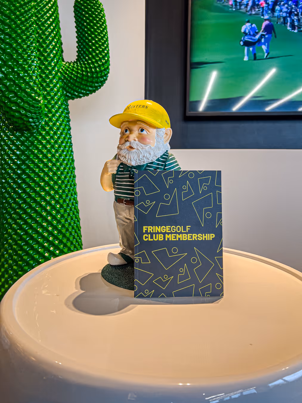 Person preparing to hit a golf ball indoors with a golf bag labeled 'FRINGEGOLF' on artificial turf and a golf simulation screen in the background.