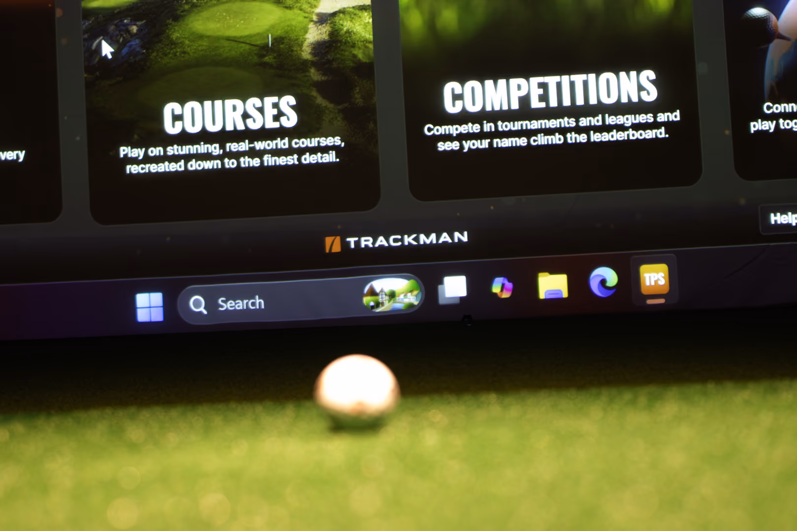 Close-up of a golf ball on turf with a screen showing Trackman golf courses and competitions.