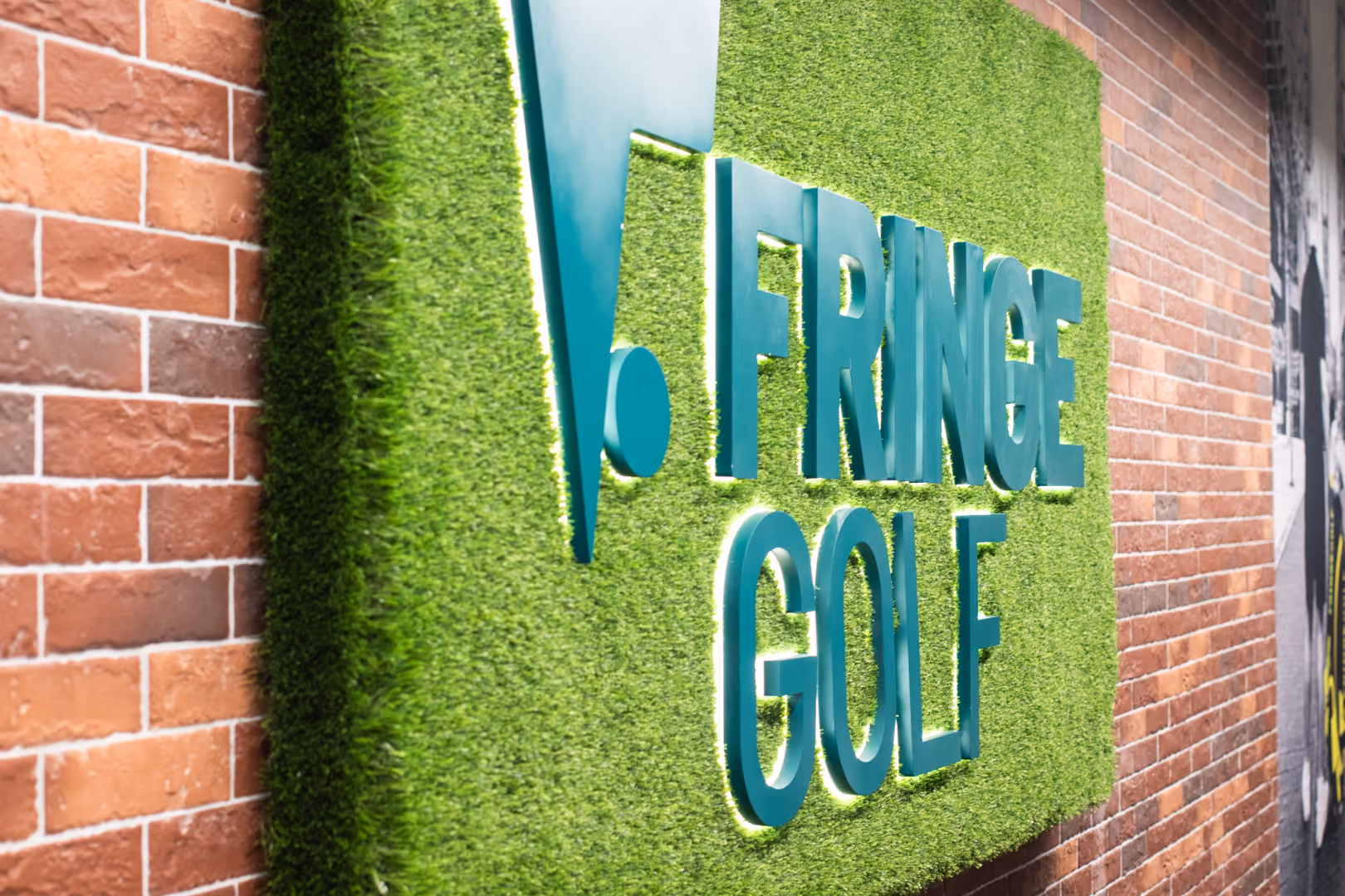 Person preparing to hit a golf ball indoors with a golf bag labeled 'FRINGEGOLF' on artificial turf and a golf simulation screen in the background.
