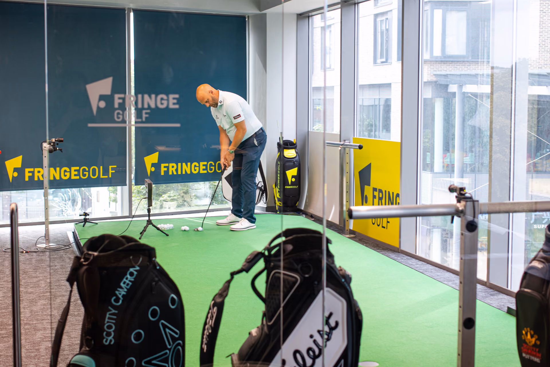 Man putting on an indoor green with golf balls and a camera set up, in a room with large windows showing city buildings.