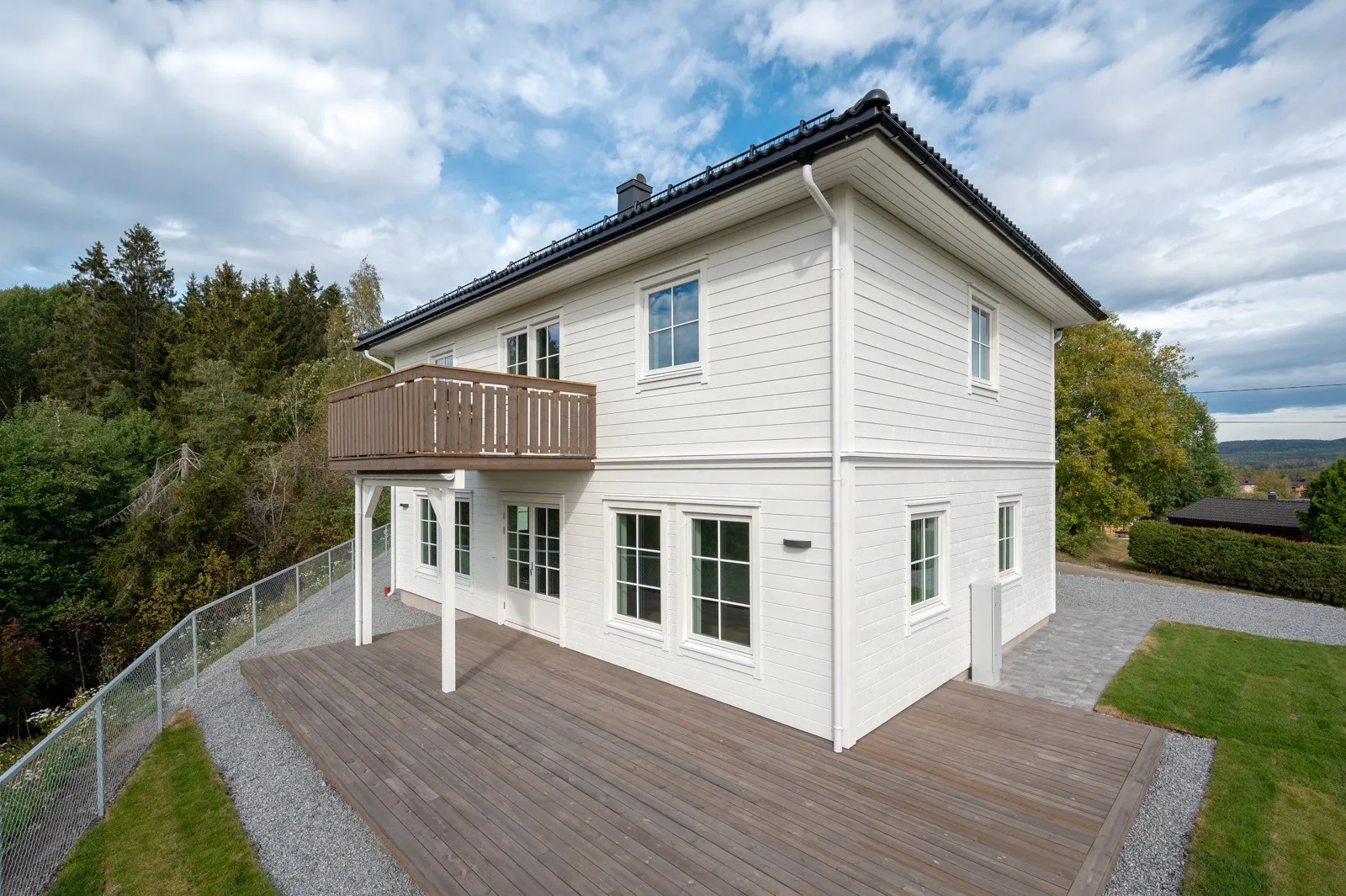 Et toetasjes hvitt hus med grått tak og trebalkong i andre etasje. Huset har store vinduer og en romslig treterrasse. Det er omgitt av frodige grøntområder, inkludert trær, med en overskyet himmel over hodet.