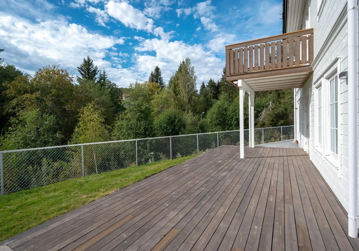 Et romslig treterrasse festet til et hvitt hus har utsikt over en frodig grønn skog. Over er det en trebalkong. Himmelen er delvis overskyet, noe som bidrar til den naturskjønne og rolige utendørsomgivelsen.