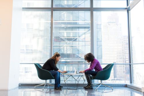 Zwei Frauen sitzen in einem modernen Bürogebäude an einem kleinen Tisch und arbeiten konzentriert an Laptops.
