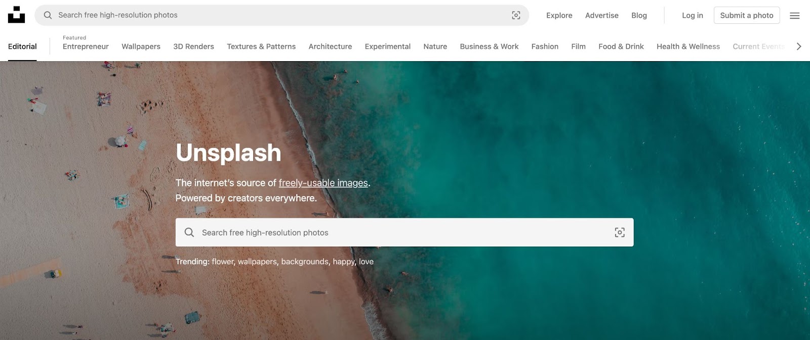 Unplash homepage, background is a drone-shot of a a beach with turquoise waters and a few people sitting on towels in the sand. middle of page shows a search bar for photos. 
