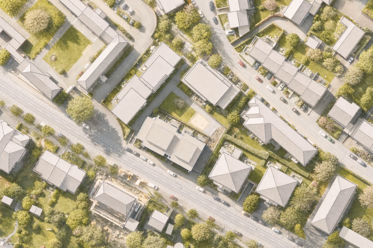 Aerial view of a residential neighborhood with multiple houses, green lawns, trees, driveways, and cars parked along streets and in driveways on a sunny day.