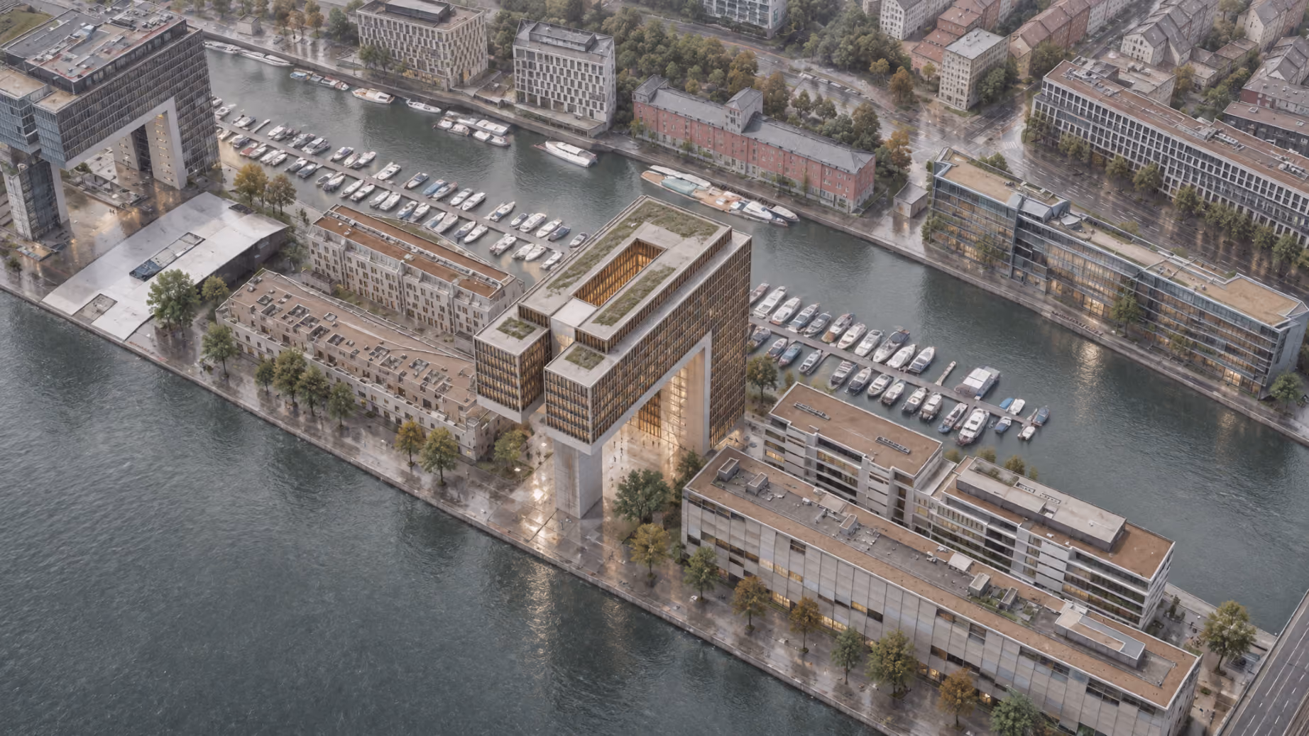 Aerial view of a waterfront urban area featuring modern buildings including two large structures with central open spaces, marinas with docked boats, tree-lined walkways, and streets with cars and greenery in the background.