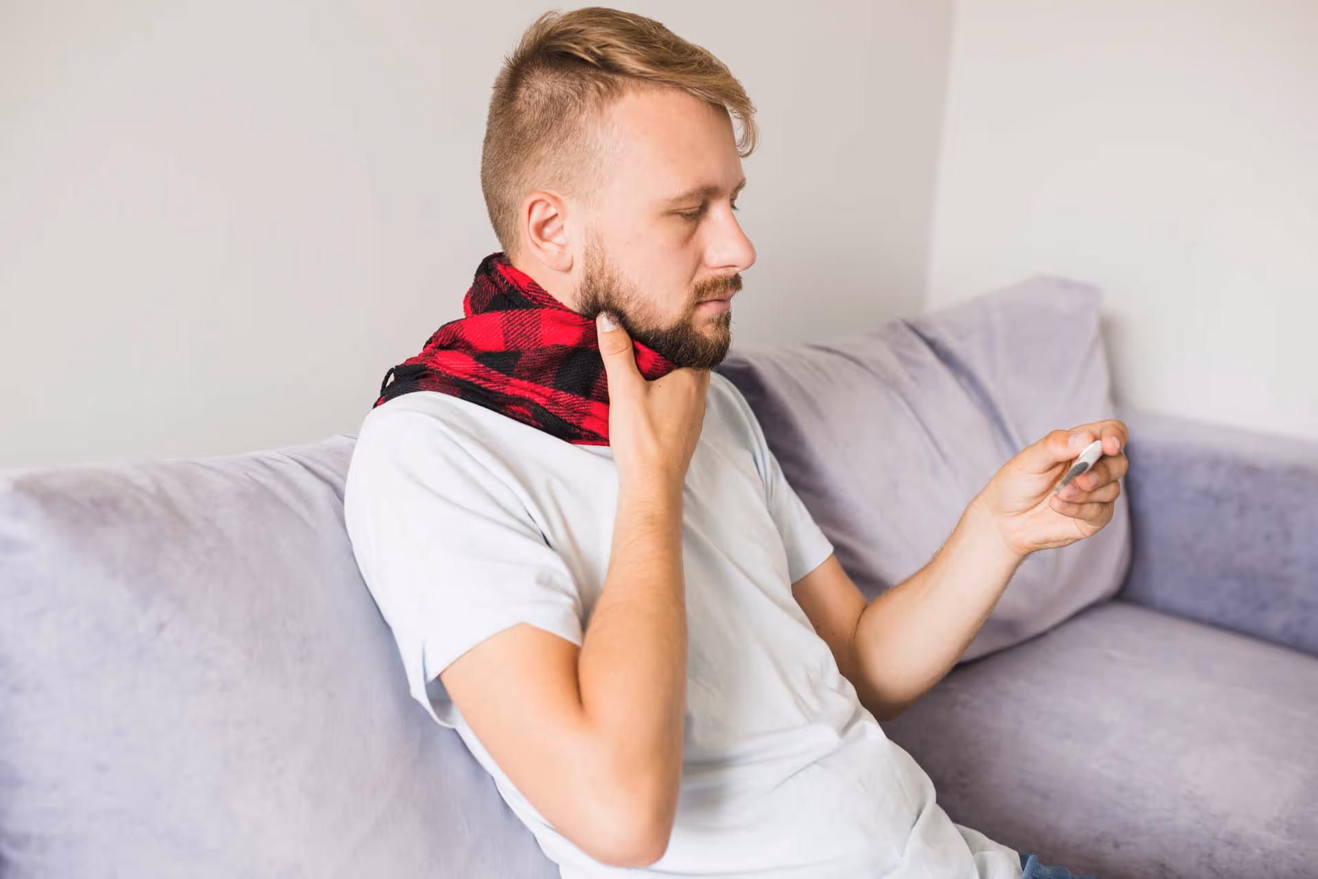 Man sitting on a couch checking a thermometer, touching his throat, with a red scarf around his neck.