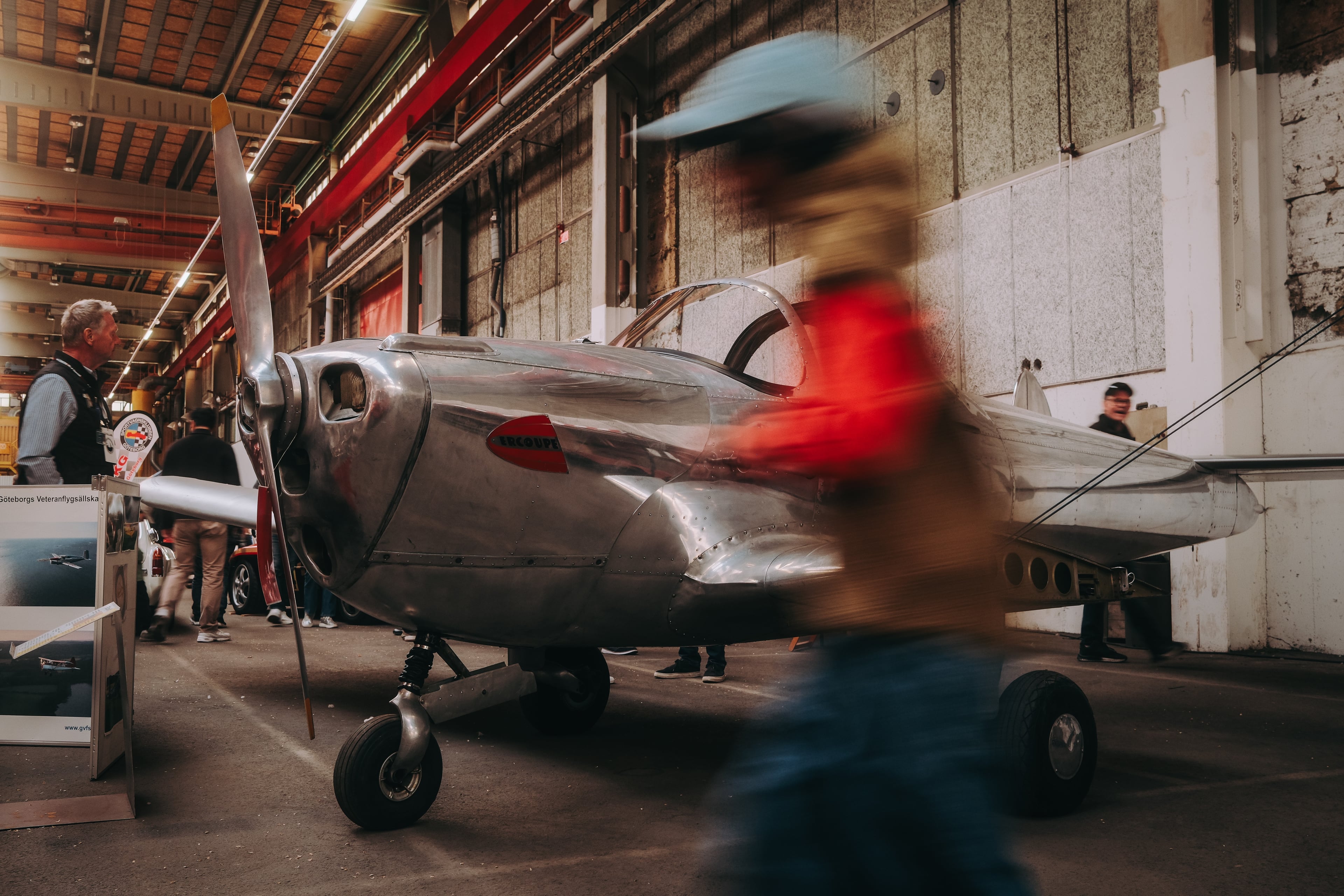 En liten blank metallpropellerflygplan visar sin främre sektion med propellern i en inomhusmiljö med flera personer runt omkring.