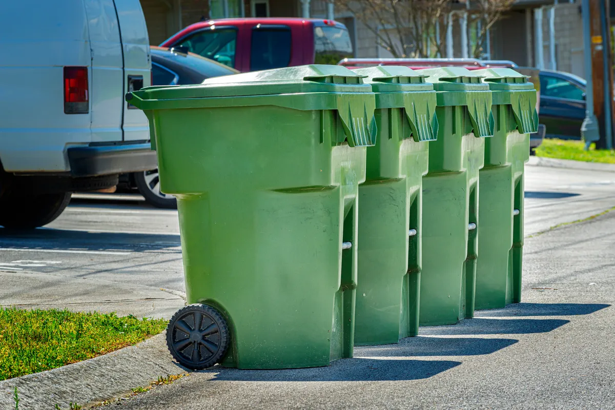 A row of green trash cans lined up neatly against a backdrop of grass and trees