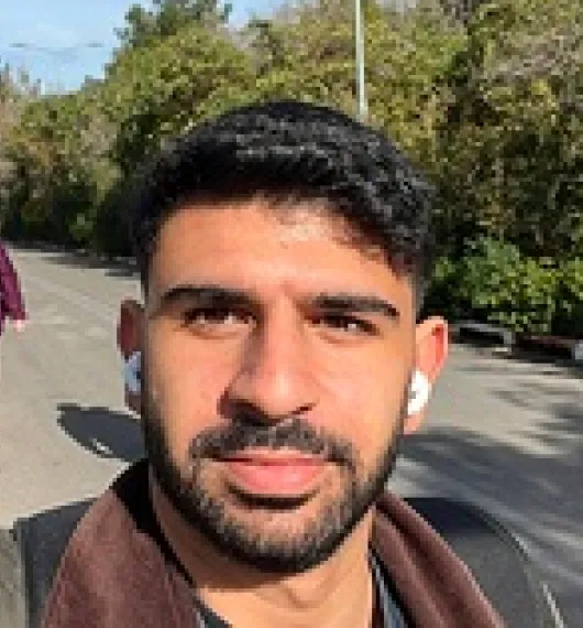 Close-up of a young man with short black hair and a beard wearing white earbuds outdoors on a sunny day.