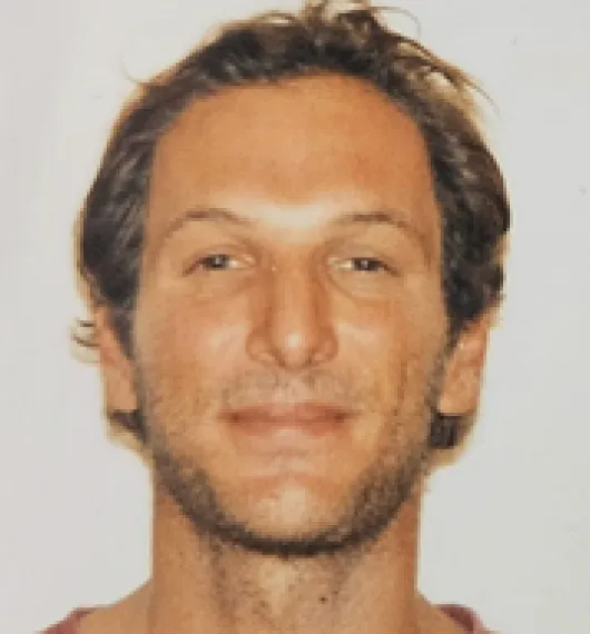 Close-up of a smiling man with short brown hair and light facial stubble against a plain background.