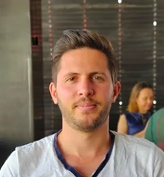 Young man with short brown hair and light beard wearing a white shirt seated indoors with a blurred background showing another person.
