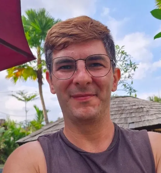 Man wearing glasses and a sleeveless black shirt outdoors with tropical plants and a thatched roof in the background.