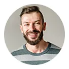 Smiling man with short brown hair and beard wearing a gray striped shirt against a light background.