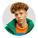 Young man with curly brown hair wearing a green jacket and orange sweater, looking directly at the camera against a plain background.