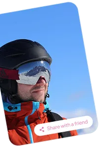 Man wearing a black helmet and reflective ski goggles with snowy mountains reflected, dressed in an orange and blue jacket against a clear blue sky.